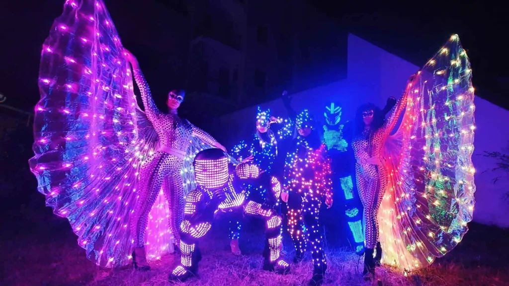Performer in costumi luminosi si esibiscono su un palco buio con grandi ali LED multicolore e decorazioni brillanti
